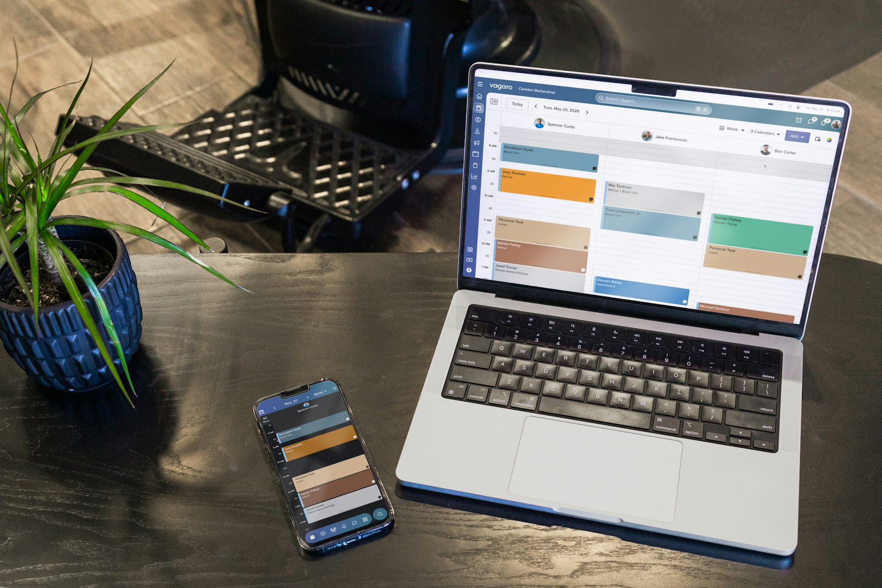 A laptop and a phone showing a scheduling calendar on a black desk beside a plant