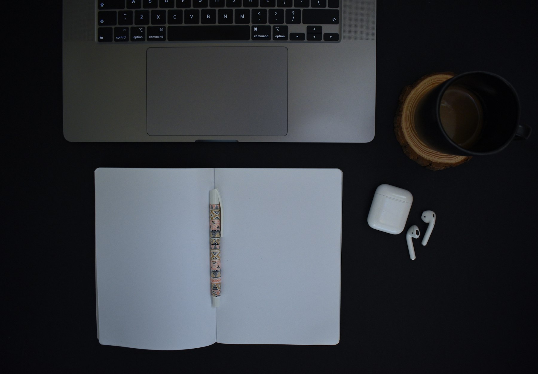 A laptop, an open notebook with a pen, a coffee mug and earbuds laid out on a black desk