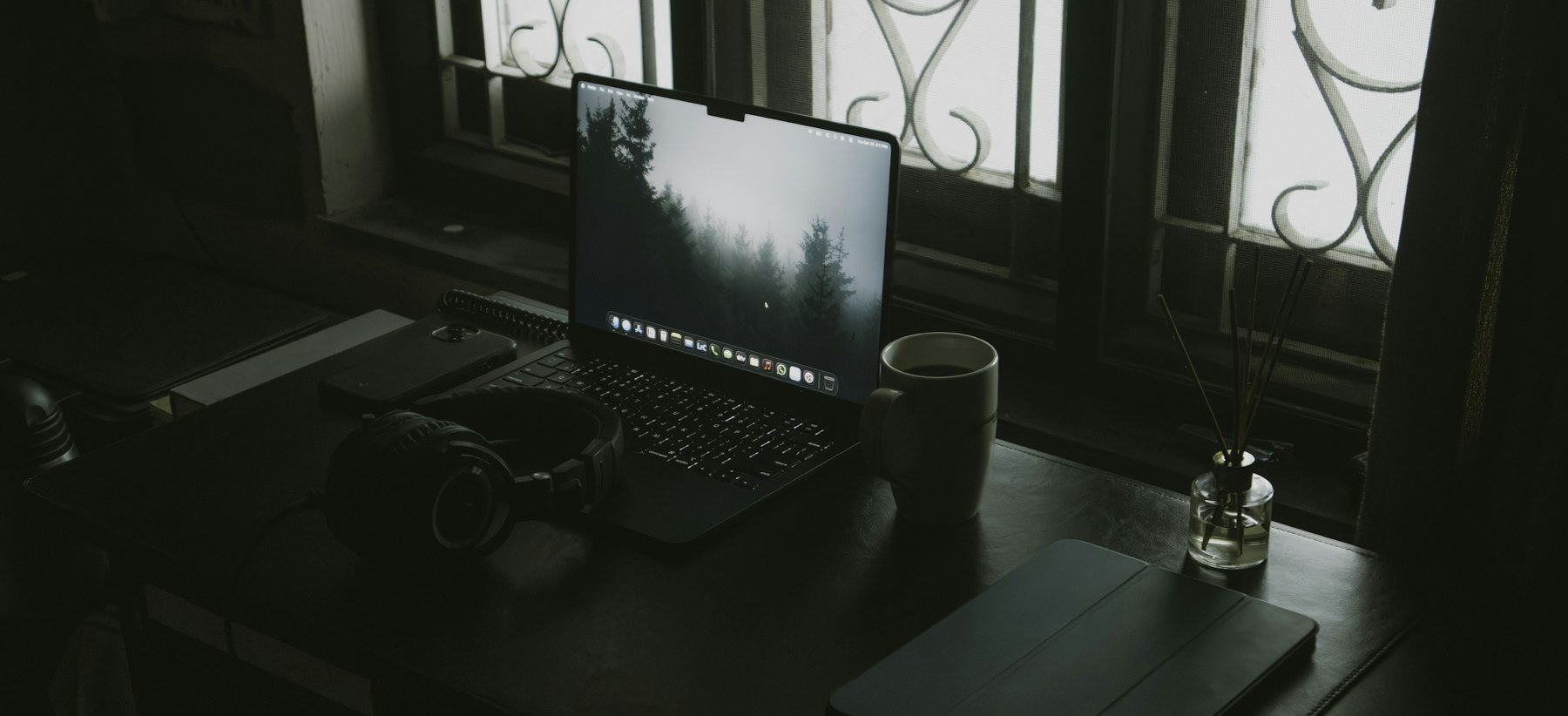 A laptop, headphones and a coffee cup on a dark desk beside a bright window