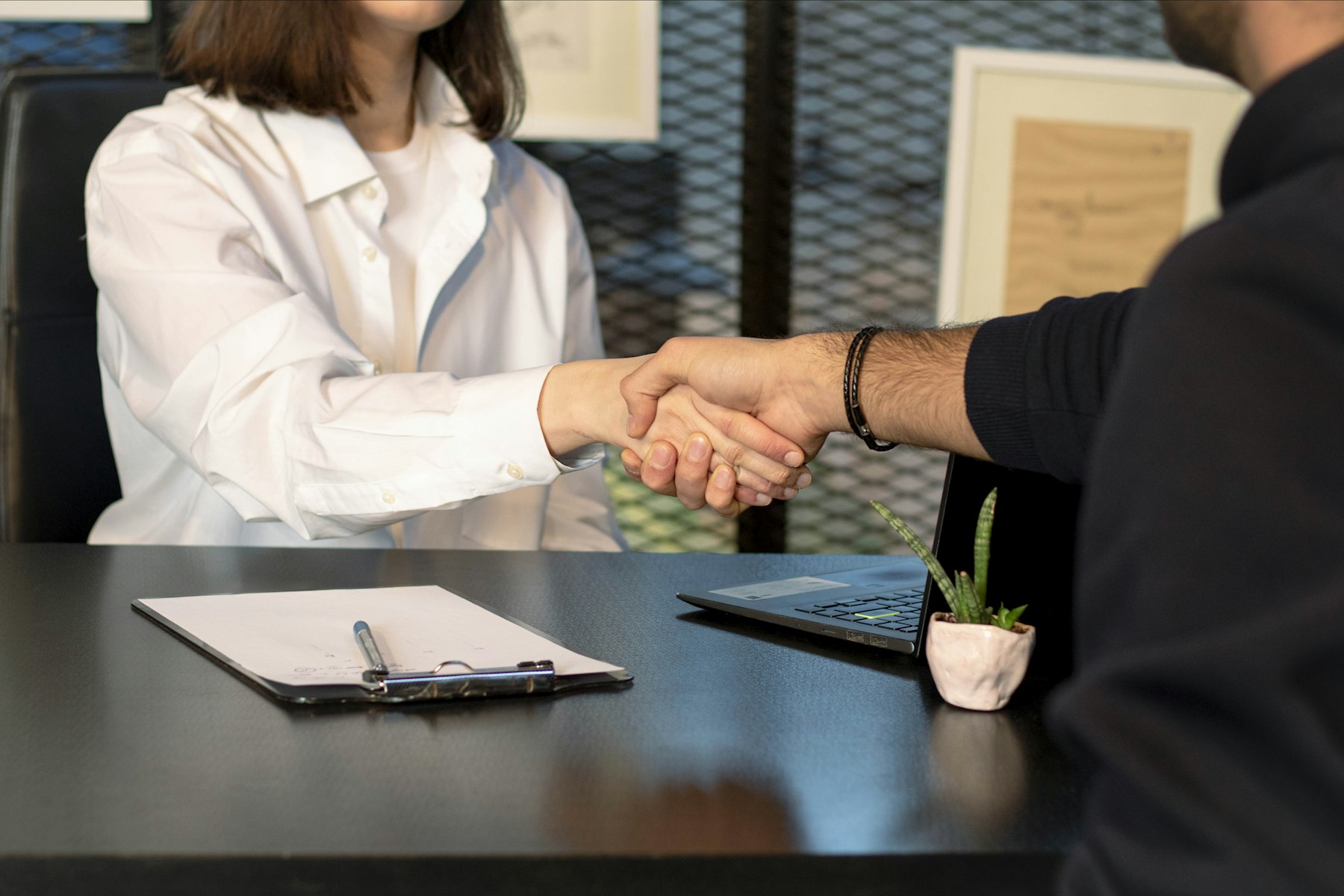A man and a woman shaking hands across a desk, a laptop and clipboard between them
