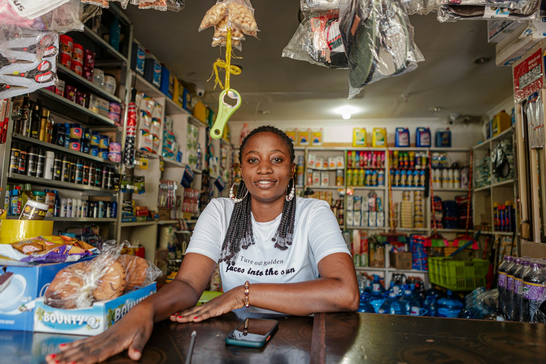 A woman behind the counter of her corner shop, shelves stocked behind her