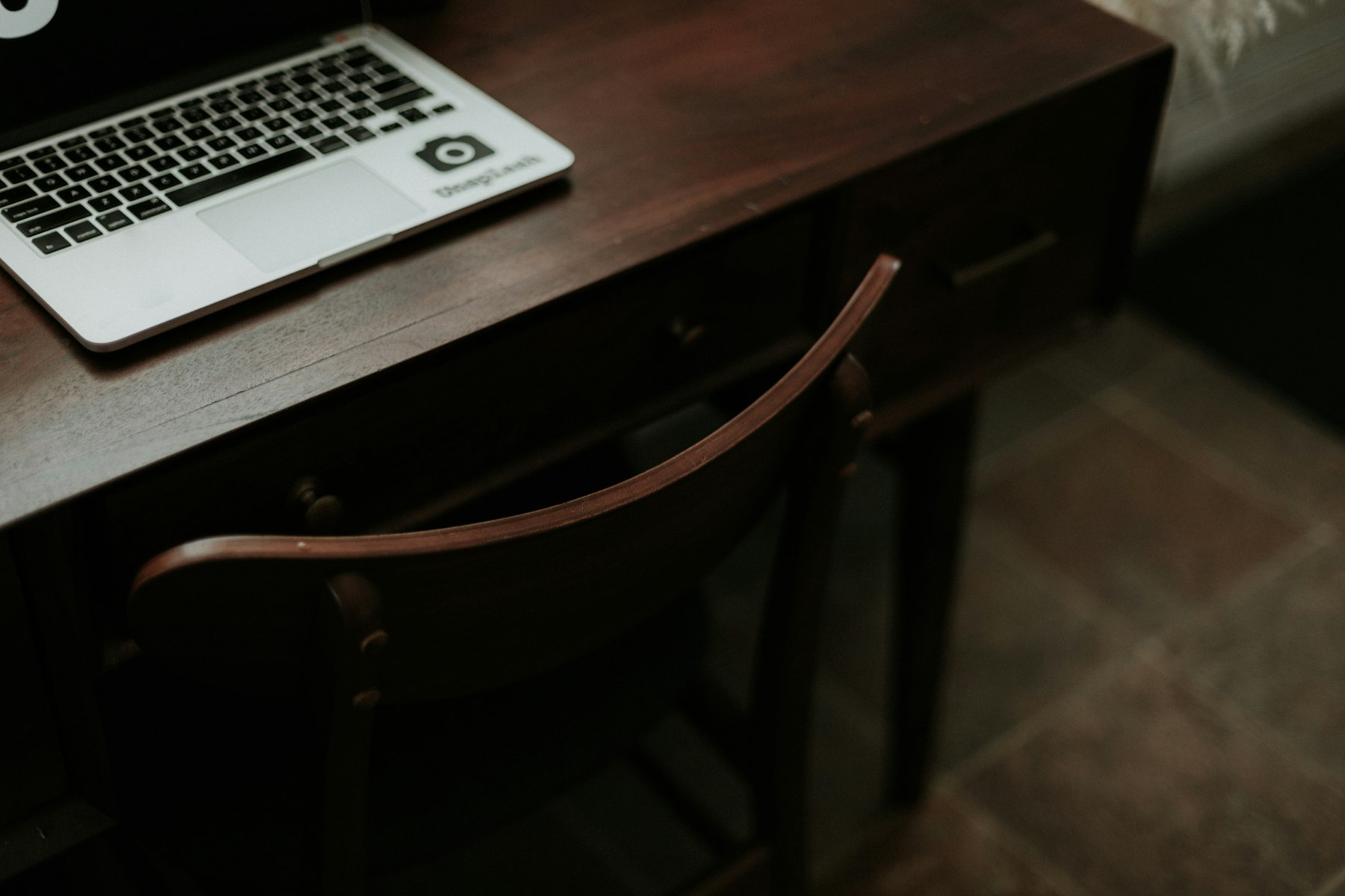 A laptop on a dark wooden desk with a wooden chair pulled out
